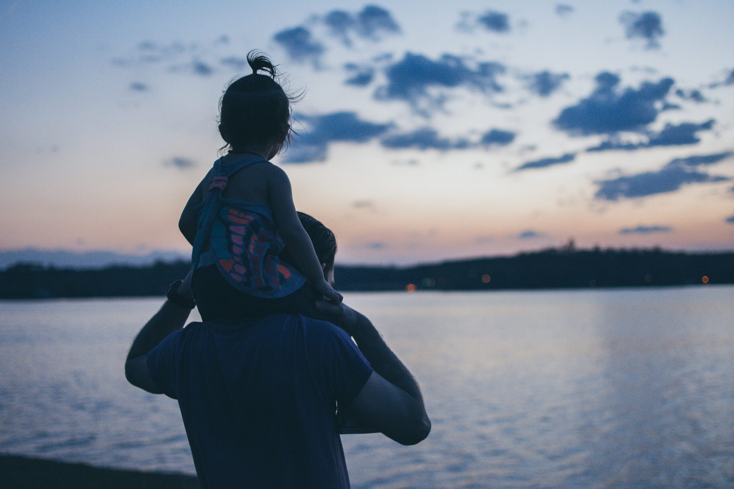 Cosa regalare alla festa del papà? bambino sulle spalle del papà al tramonto sul mare.