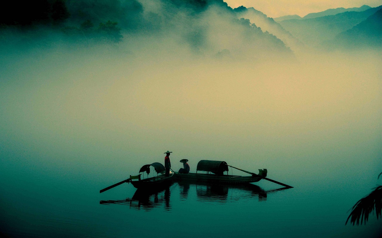 Viaggio in Cina 2024. Silhouette di due pescatori su un lago, avvolti dalla nebbia, con montagna sullo sfondo.
